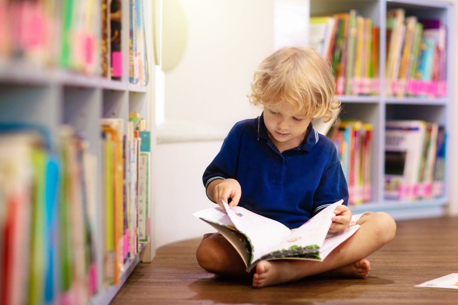 A child sitting on the floor in a library, looking down at an open book with hands turning the pages.