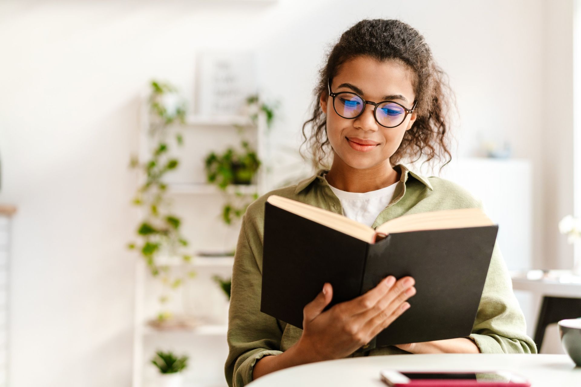 Woman reading a black book at a desk in a bright room with plants.
