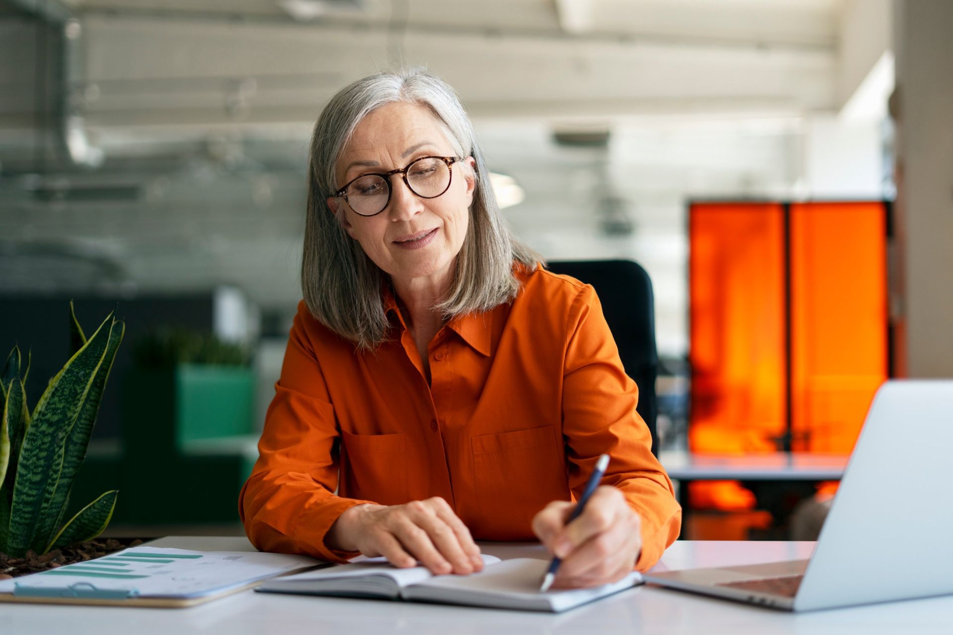 A person wearing glasses and an orange shirt sits at a desk, writing in a notebook next to a laptop and a houseplant.