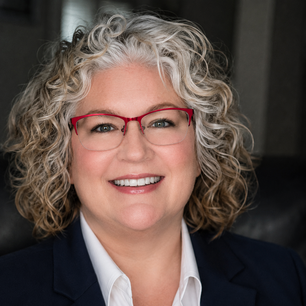 A person with curly, shoulder-length blonde hair and red-framed glasses, smiling while wearing a black high-neck top.