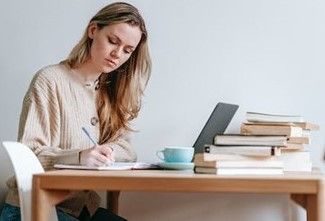 A person sitting at a wooden table, writing in a notebook next to a laptop, a blue cup, and a stack of books.