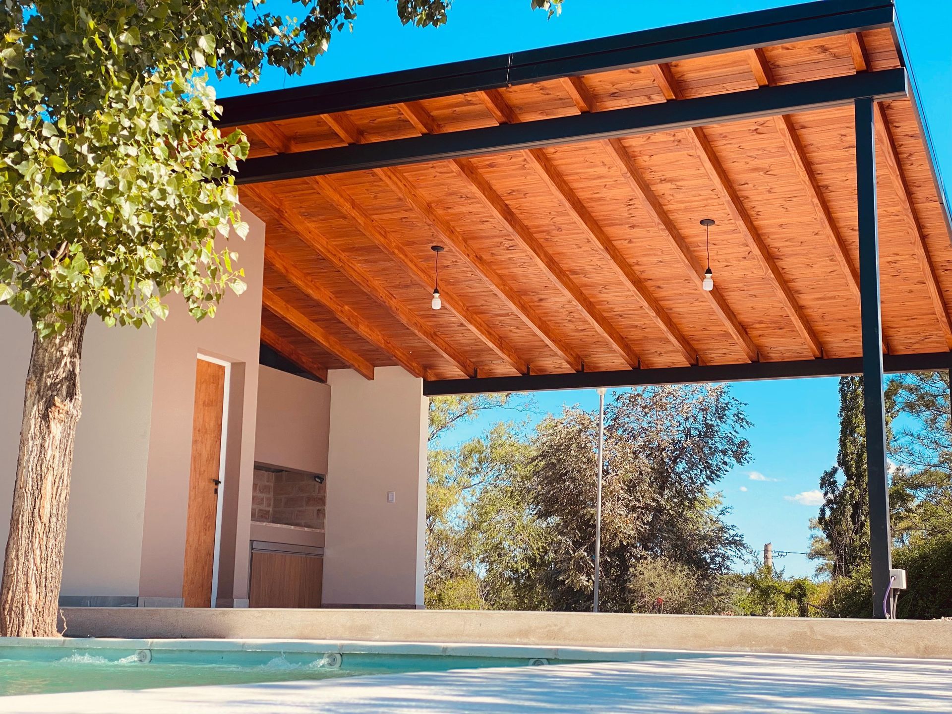 Patio junto a la piscina con techo de madera, paredes blancas y vigas negras.
