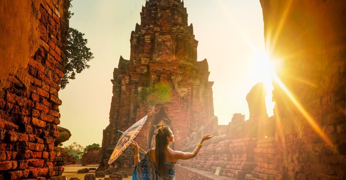 Woman in traditional dress, holding parasol, poses at ancient temple ruins during sunset.