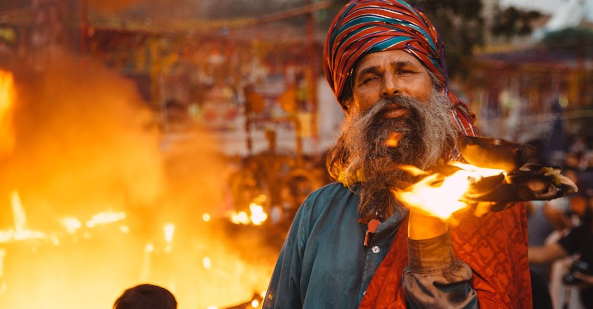 Man with beard and turban holding flaming object, set against fire and crowd.
