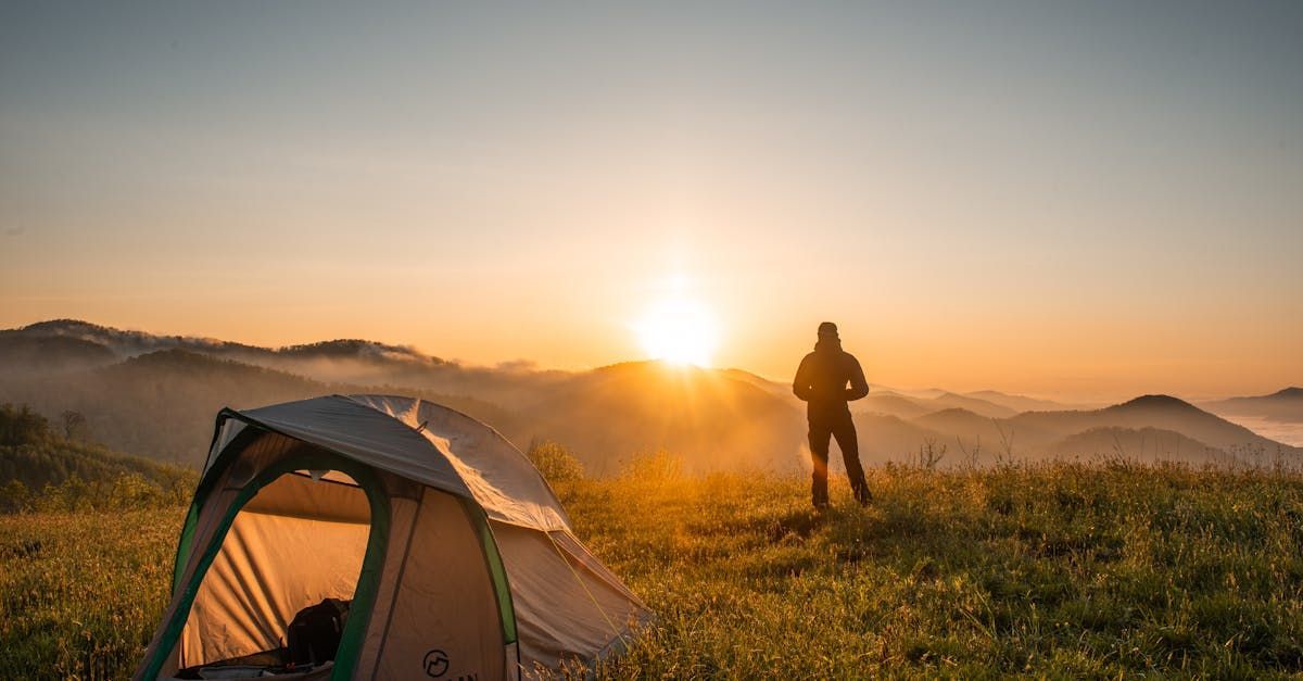Man standing on a hilltop at sunrise near a tent. Warm light on the landscape.