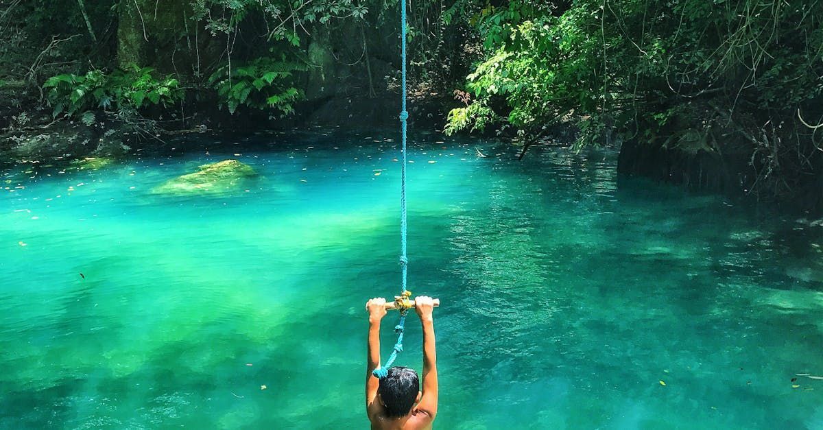 Person swinging on a rope into turquoise water surrounded by lush greenery.