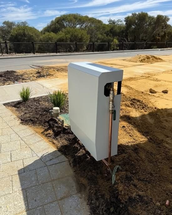White electrical enclosure with copper pipe in a dirt patch next to a brick sidewalk and street.