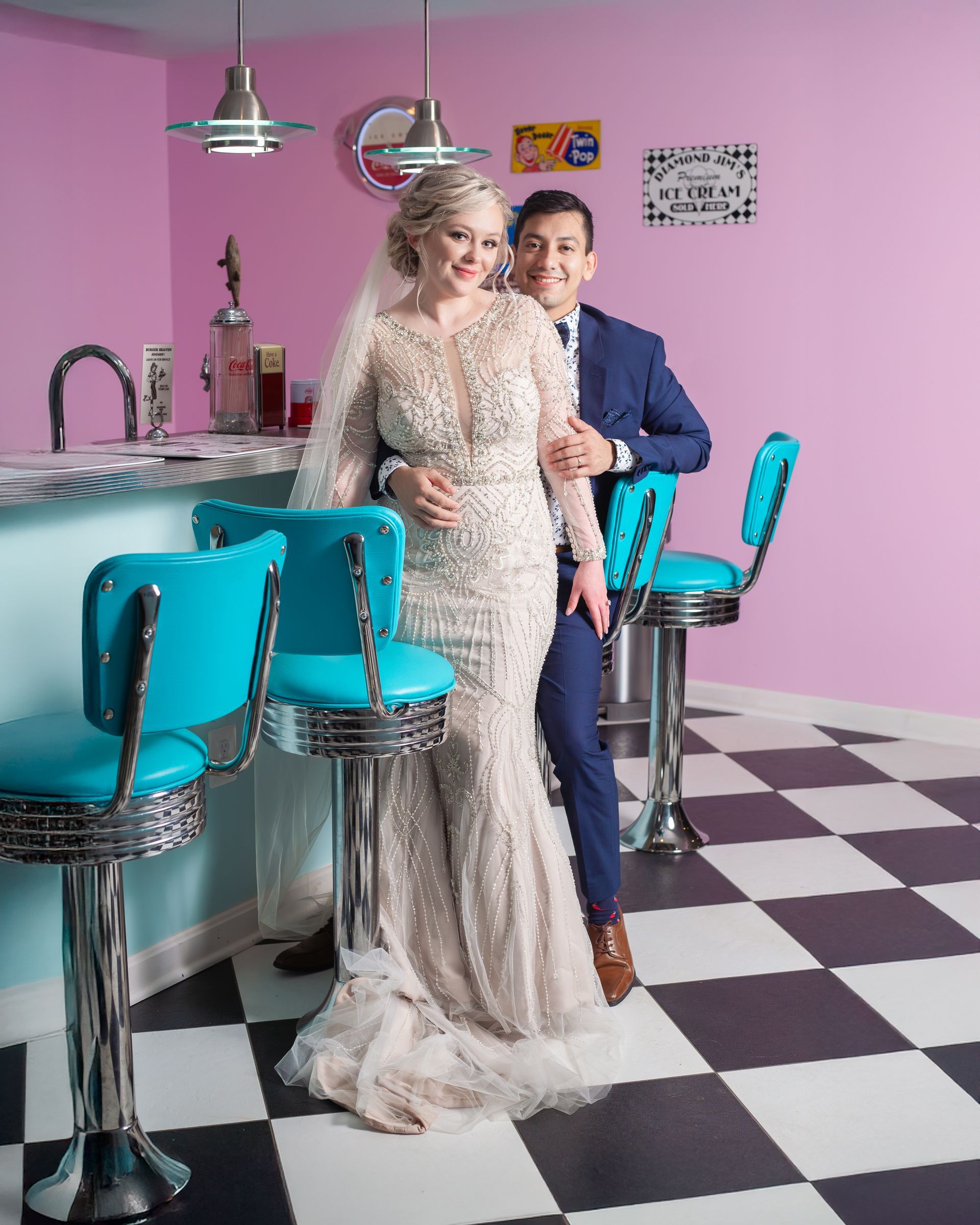 Bride and groom pose at a diner counter. Bride wears a gown, groom wears a blue suit. Pink walls, blue chairs.