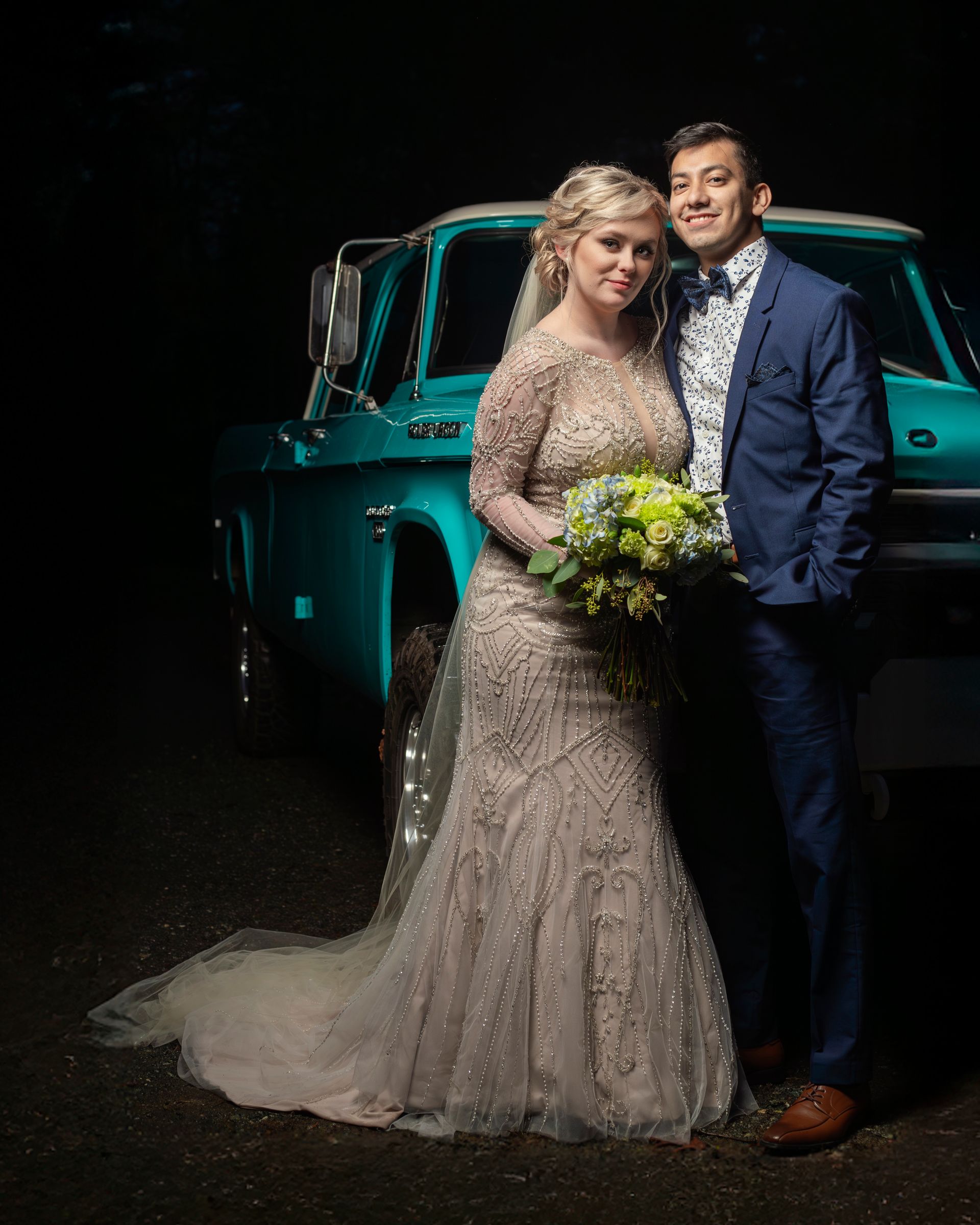 Newlyweds pose by a turquoise pickup truck. Bride in embellished gown holds bouquet; groom in blue suit.