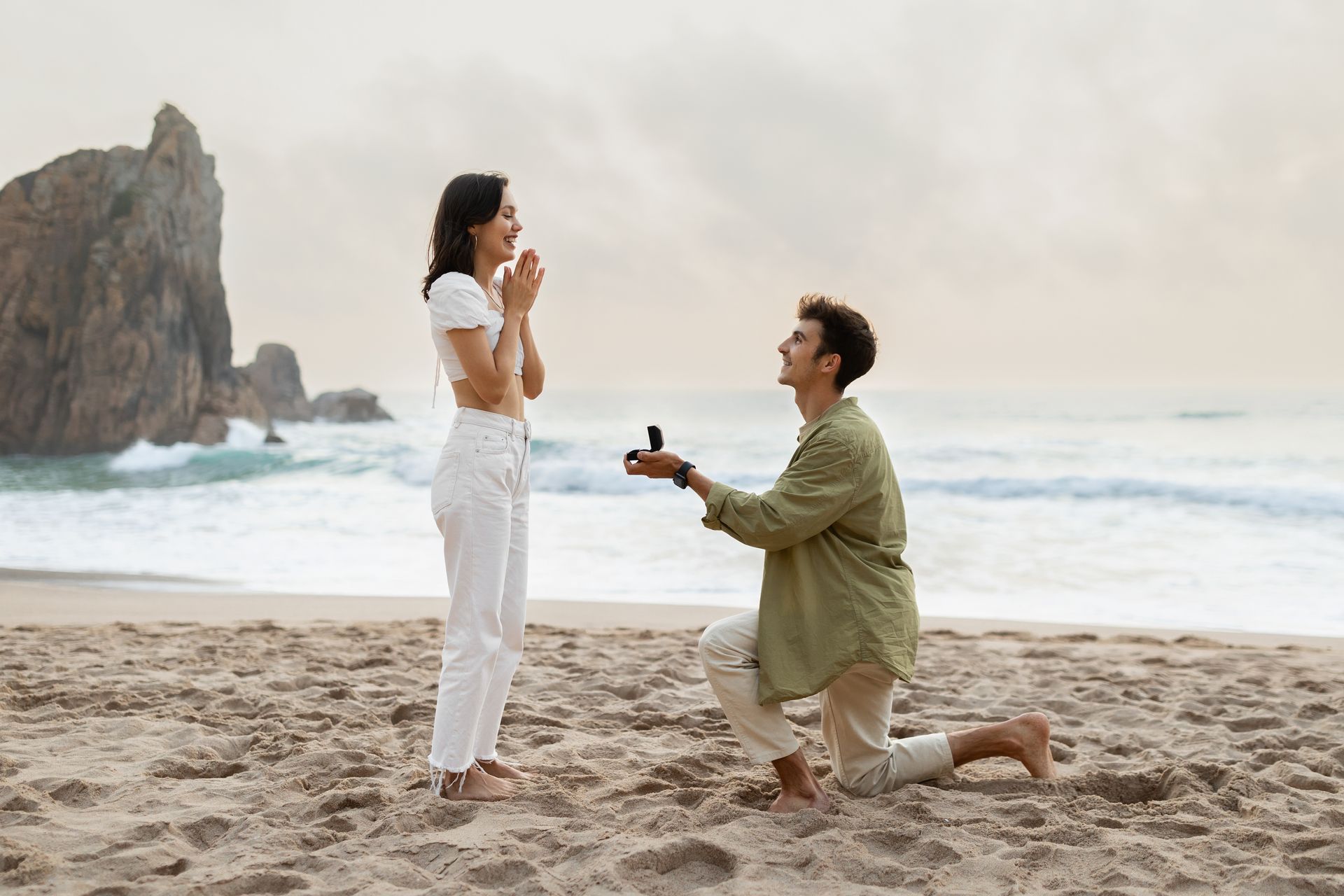 Man kneeling onng to woman; she is smiling, hands clasped near her face. Ocean and rock formations in background.