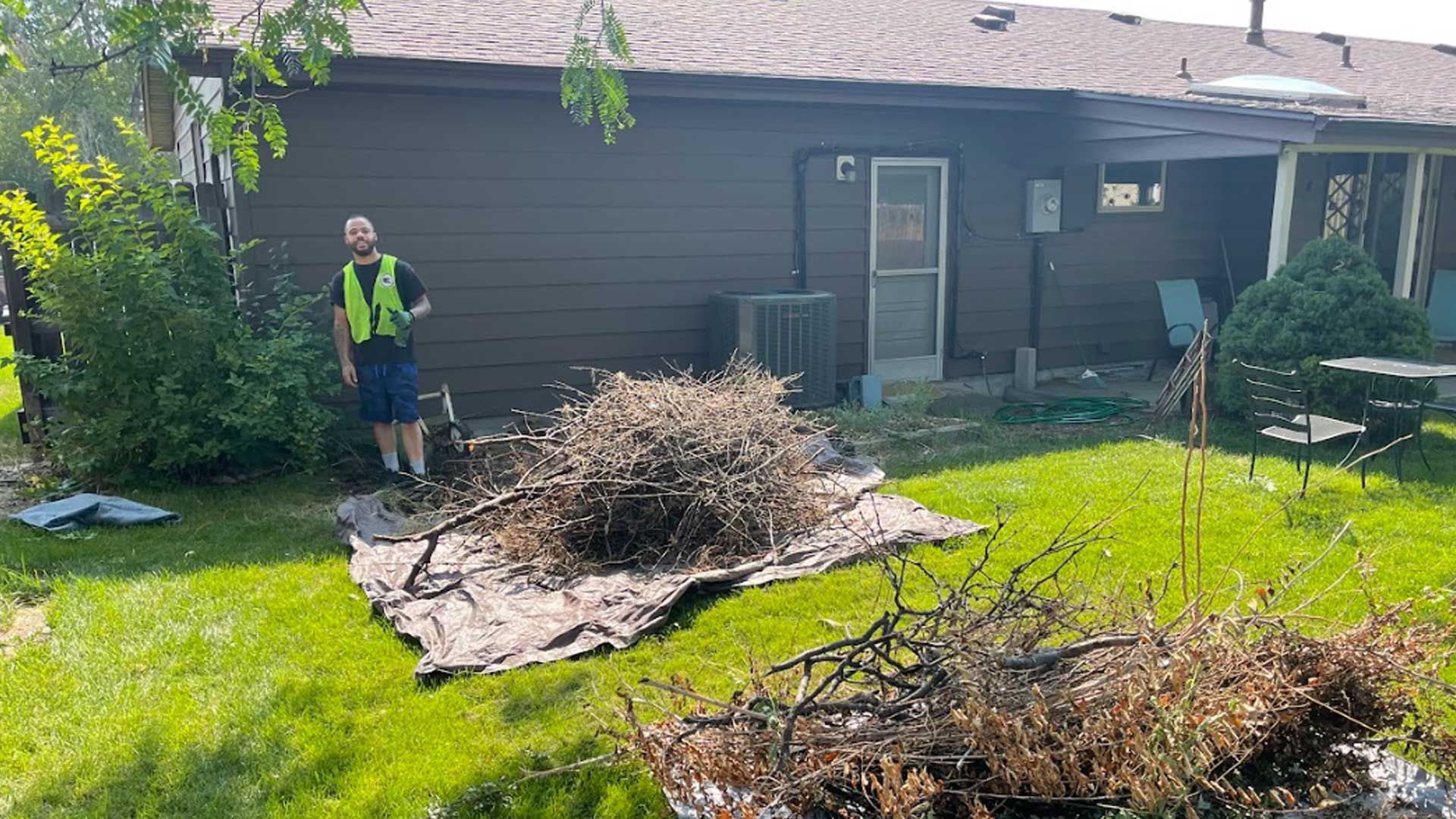 A man is standing in front of a house next to a pile of branches.
