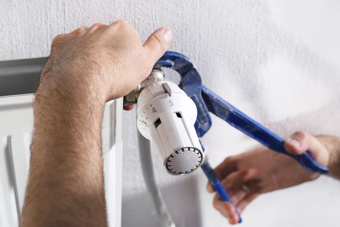 Hands using blue pliers to tighten or adjust a white radiator thermostat valve against a plain white wall.