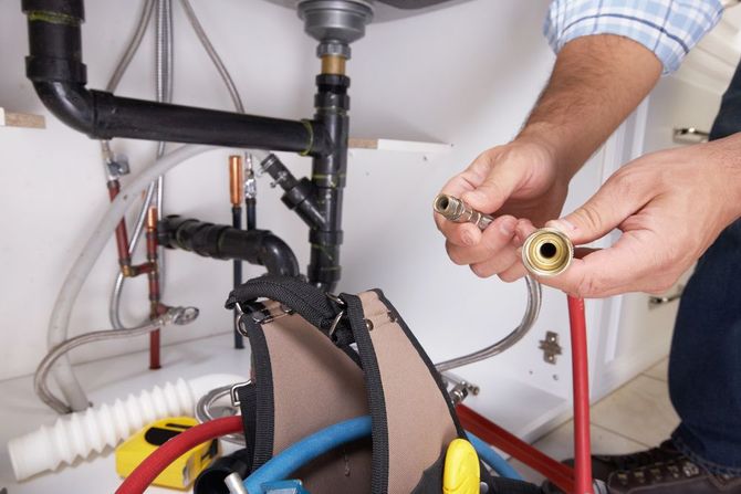 A plumber holding two metal plumbing fittings under a kitchen sink with pipes visible in the background.
