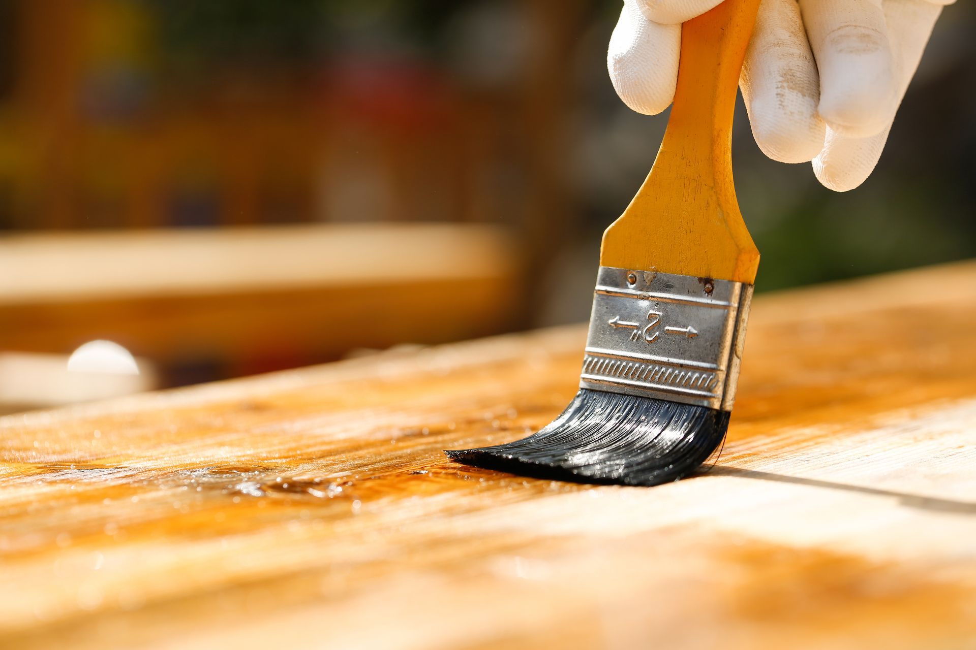 Close-up of a gloved hand applying wood stain with a paintbrush to a sunlit wooden surface.