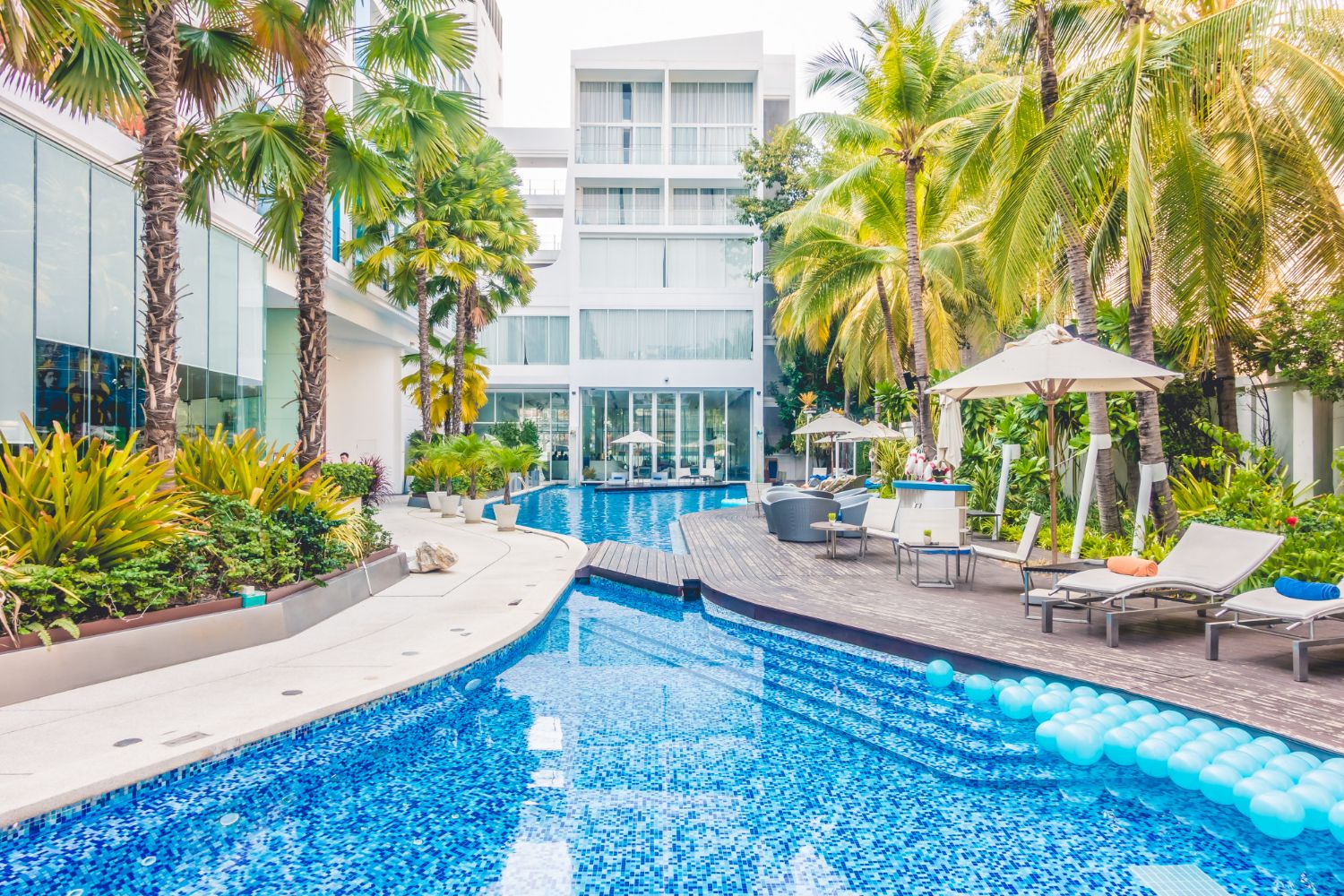 Swimming pool area at a resort with palm trees, white buildings, lounge chairs, and blue water.
