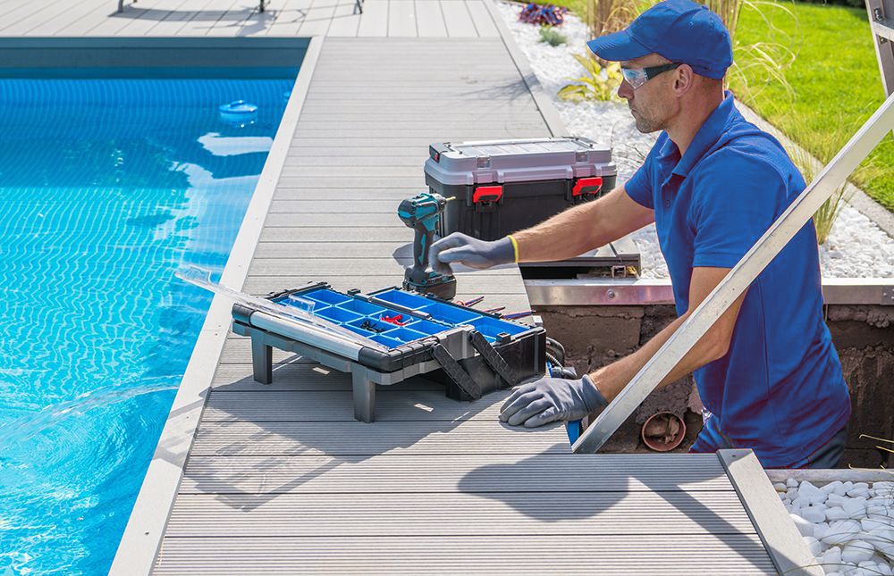 Man in blue shirt and cap repairs poolside decking. Tools and pool visible.