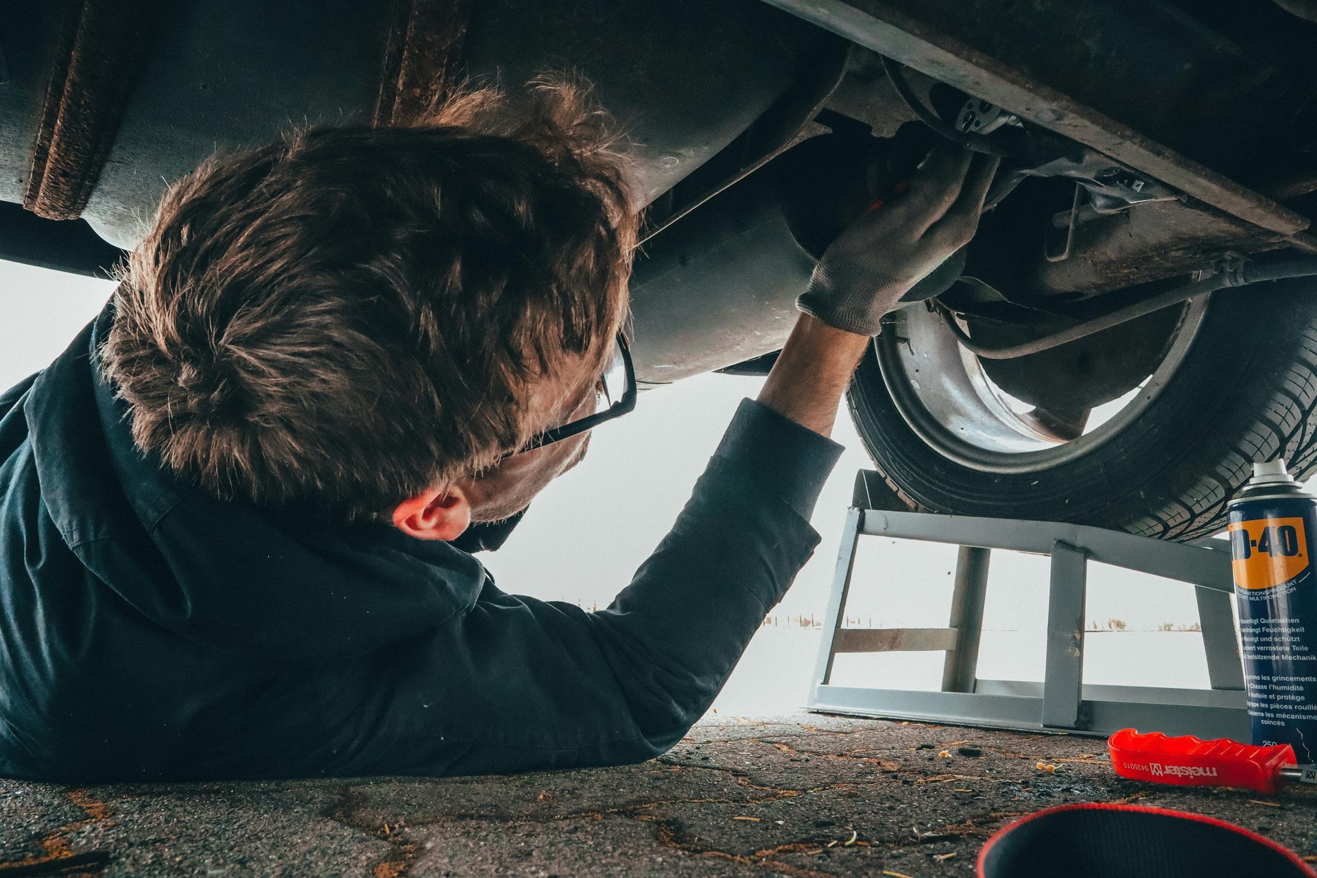 Person working on a car from below. Dark clothing, glasses, gloves. Underneath car, near tire and oil can.