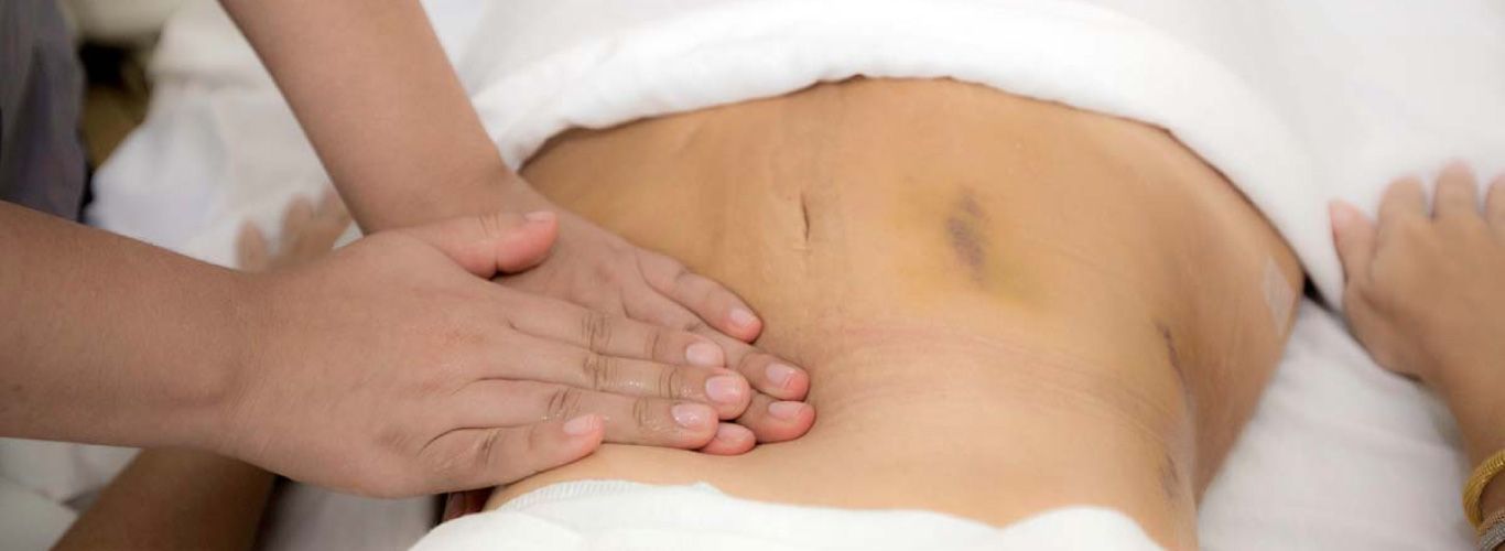 Person receiving abdominal massage on a white sheet, hands kneading stomach.
