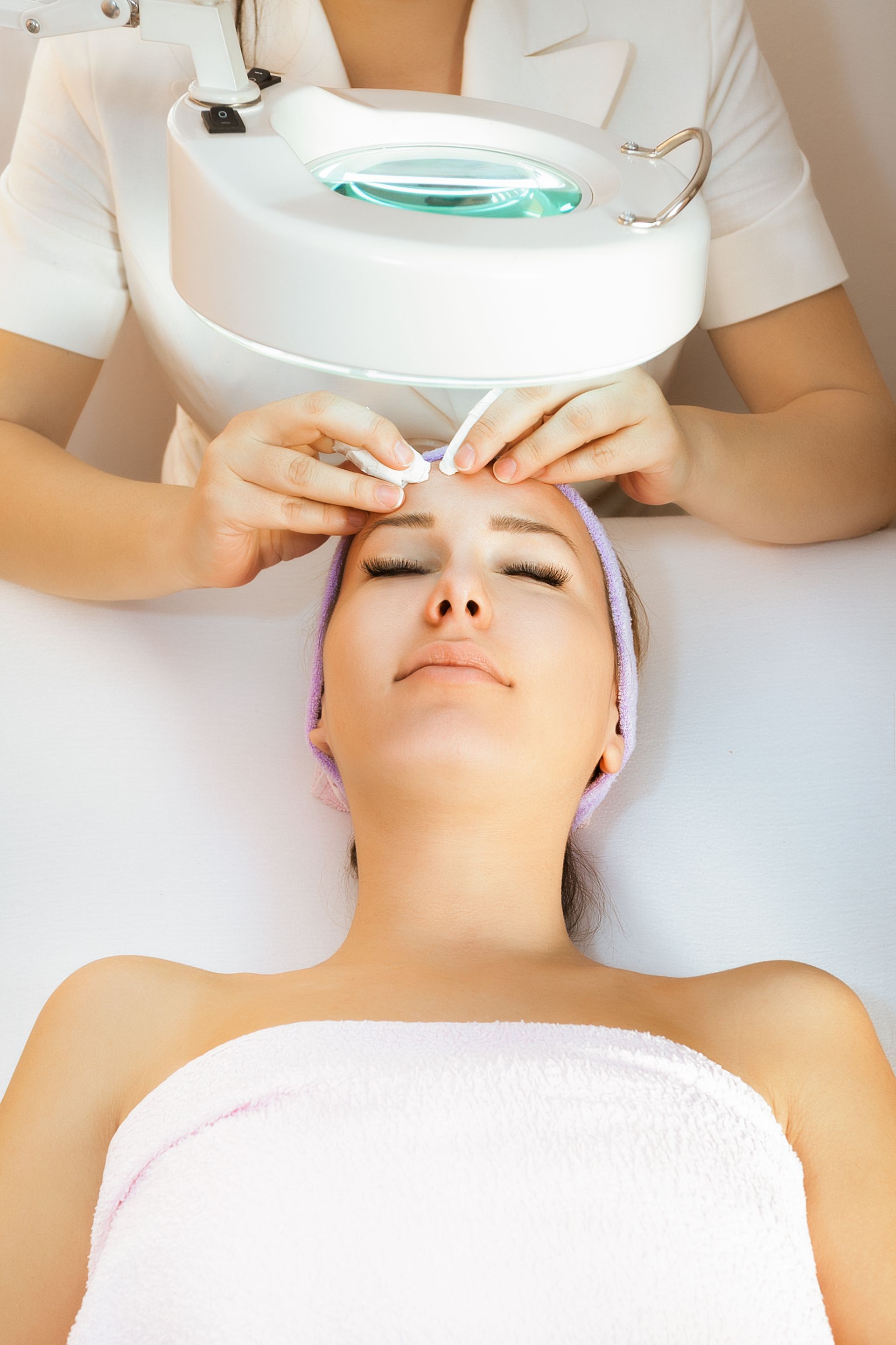 Woman receiving facial treatment at spa. Technician with magnifying lamp.