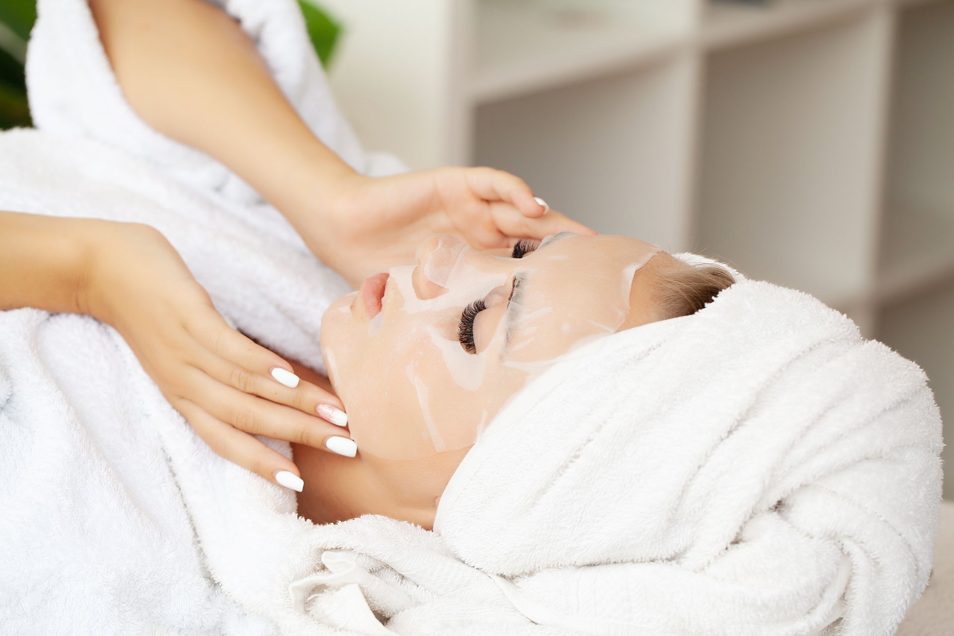 Woman in a white robe, wearing a face mask at a spa, with her hands gently on her face.