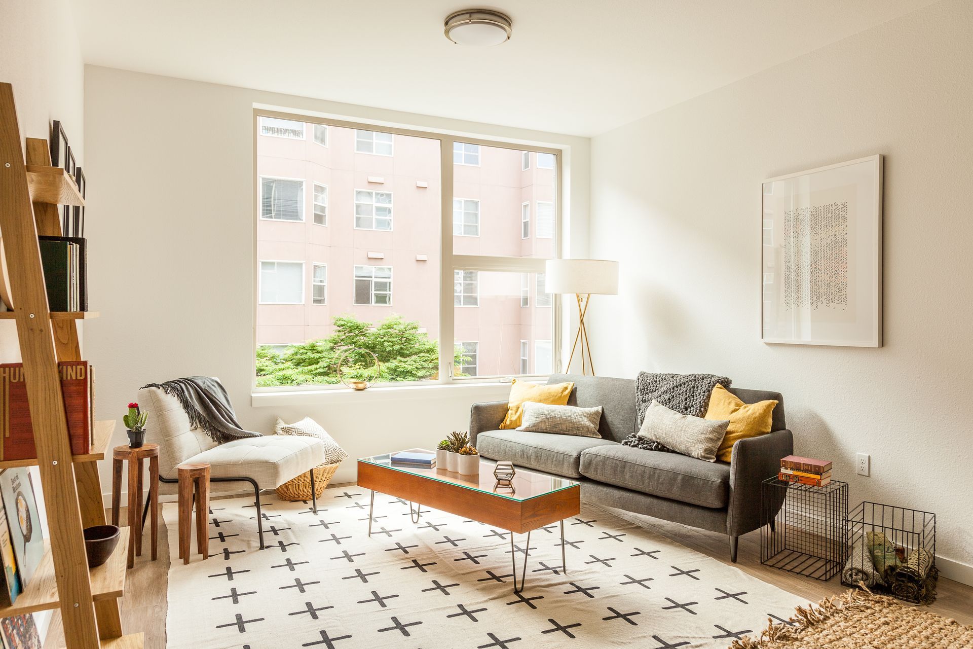 Salon confortable avec un canapé gris, un tapis à motifs et une grande fenêtre donnant sur un immeuble.