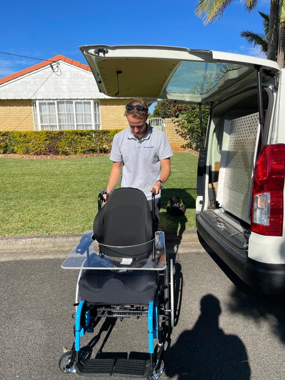 A person positions a blue wheelchair with a clear tray at the back of a parked van on a sunny day.