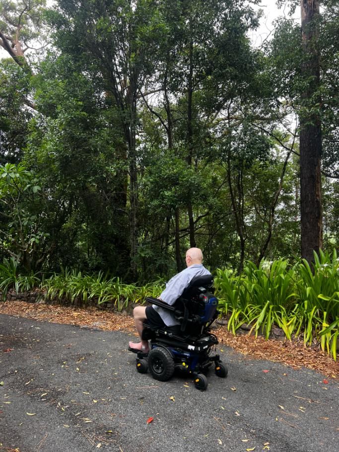 A person in a black power wheelchair drives along a paved path bordered by lush greenery and tall trees.