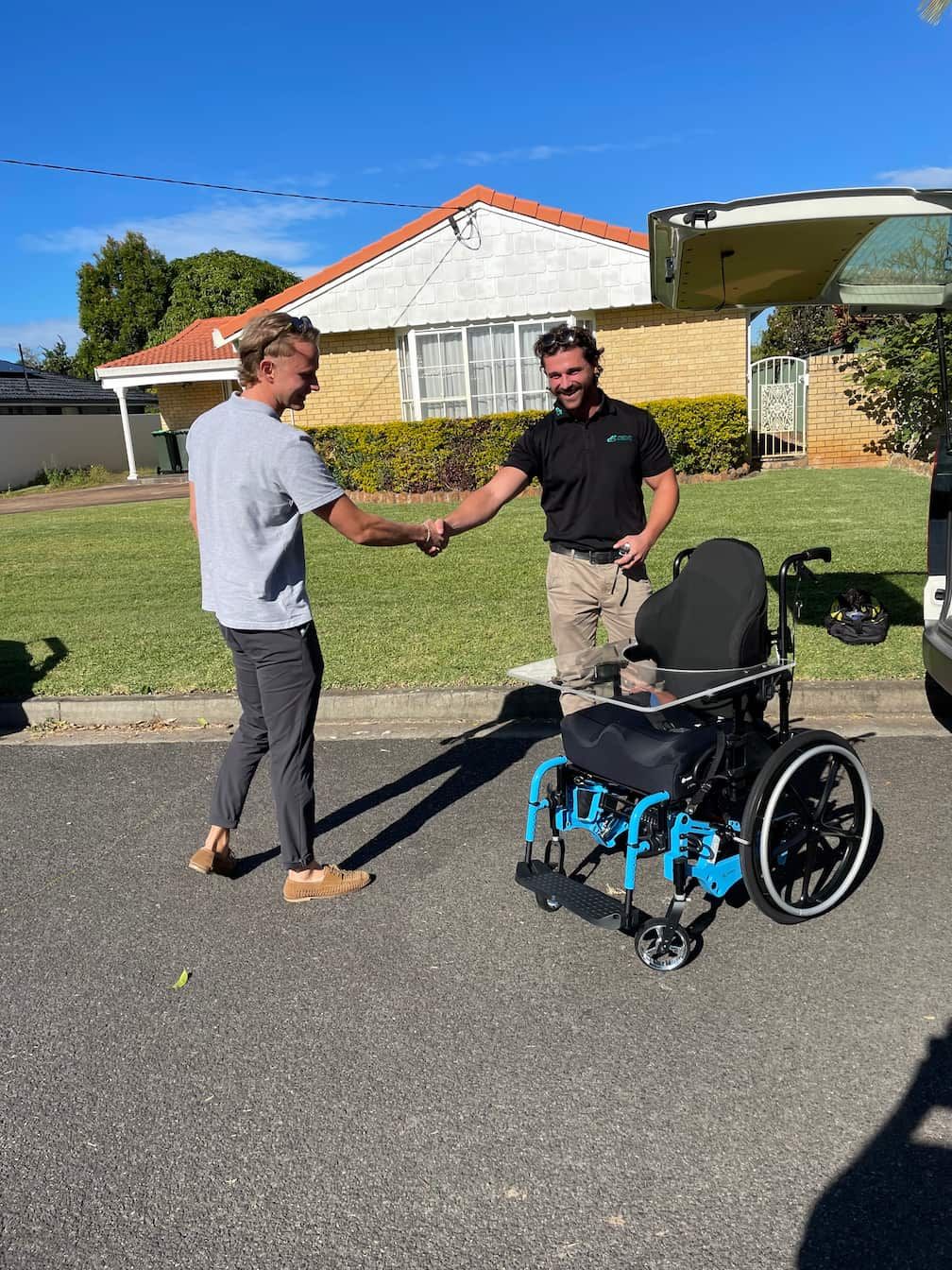 Two men are shaking hands in front of a wheelchair.