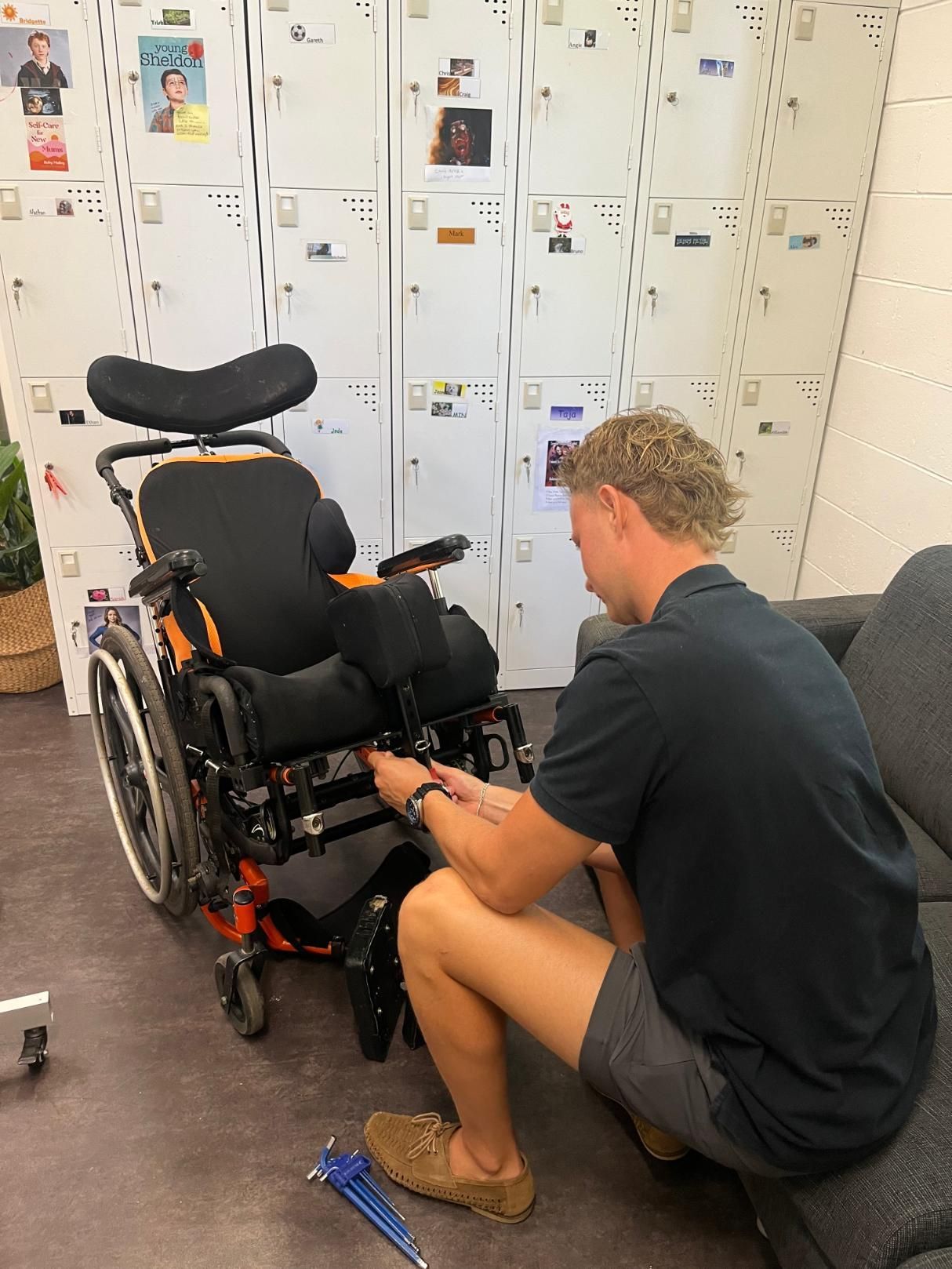 A person in a dark shirt and shorts kneels to adjust the frame of an orange and black wheelchair in an office setting.
