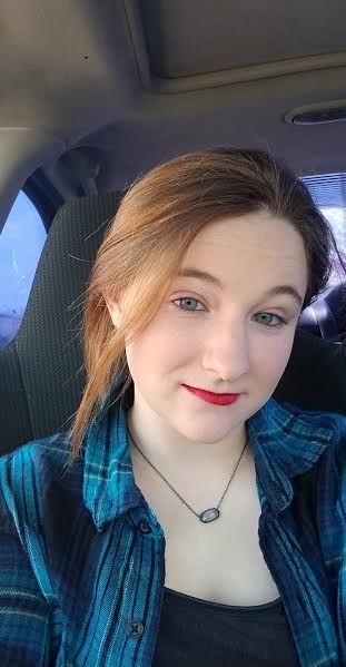 Person taking a selfie in a car, wearing a blue plaid shirt and red lipstick, with a slight smile.