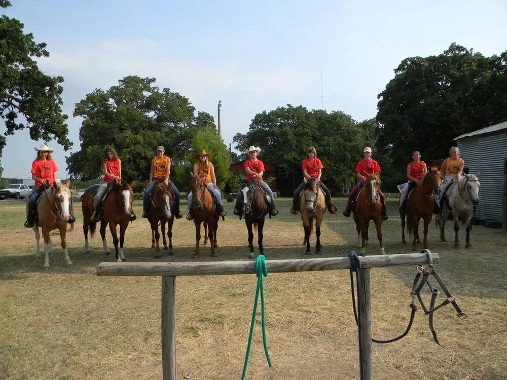 Row of horseback riders in red and orange shirts lined up in a grassy field under a blue sky