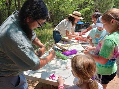 People at an outdoor table crafting with paper, scissors, and glue in a wooded area