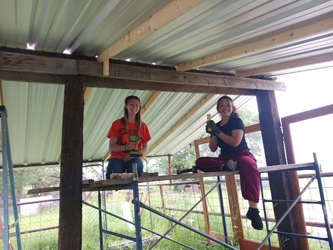 Two people sitting on a wooden platform under a metal roof, holding drinks and smiling outdoors.