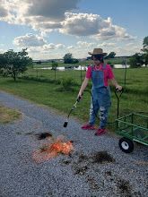 Person in a straw hat igniting a small firework near a gravel path with a cart in a grassy field