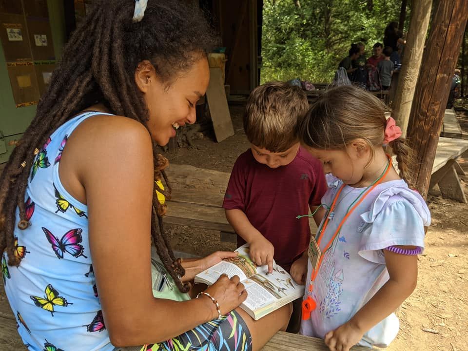 Three children gather around a table, looking at a map or book outdoors under trees.