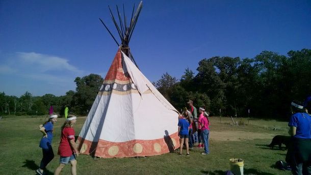 People standing around a large white teepee in a grassy field under a blue sky.