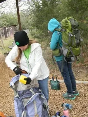 Two hikers packing gear outdoors, one adjusting a backpack in a wooded trail area.