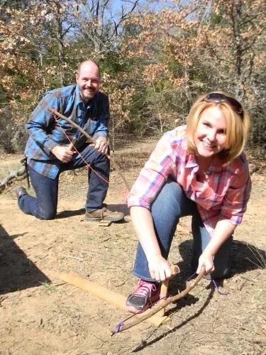 Two people kneel outdoors, using sticks to mark or measure on bare ground in a wooded area.