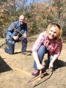 Two people kneel outdoors on a dirt path, smiling while holding a branch or small bow.