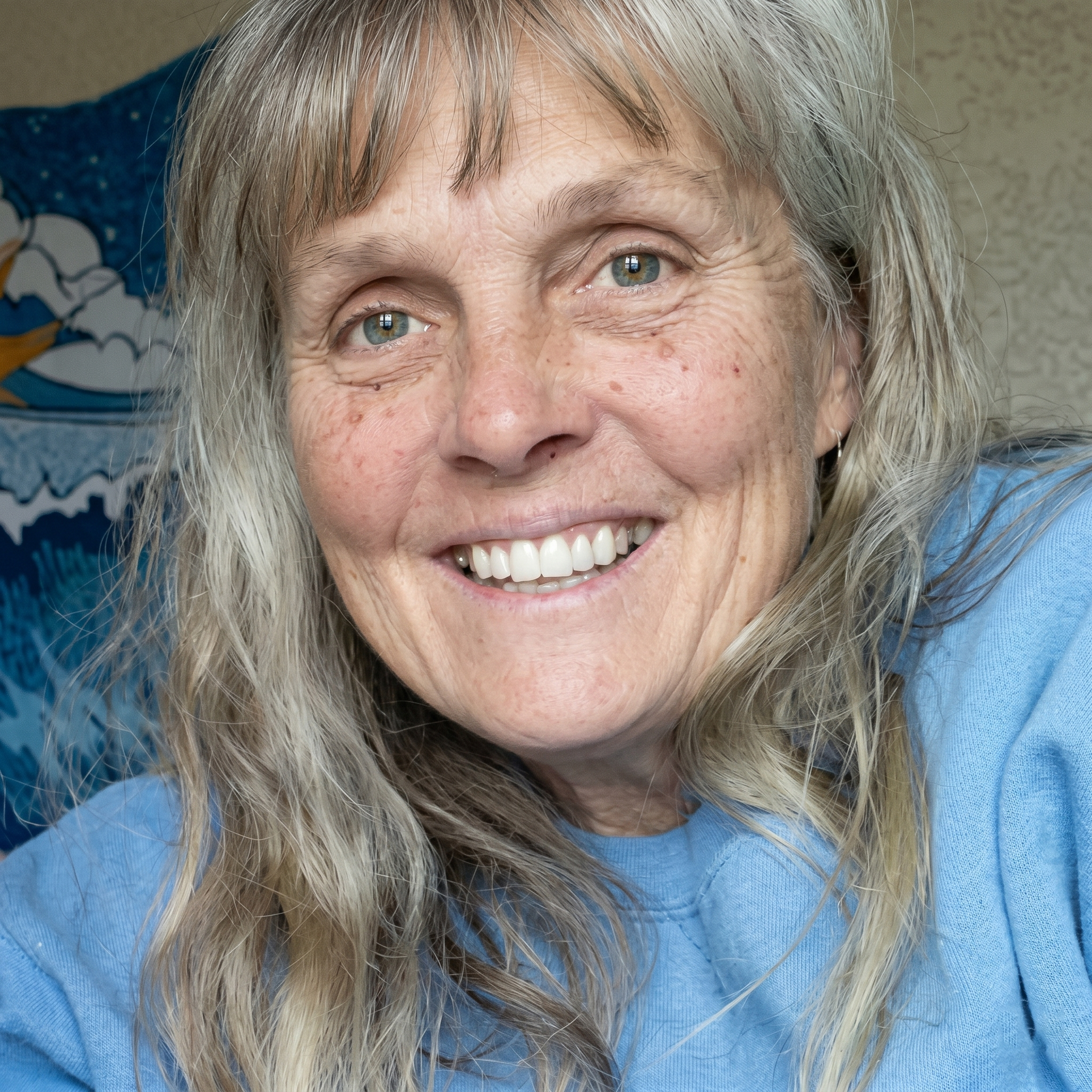 Smiling person in a blue sweater, close-up indoors, with long gray hair.