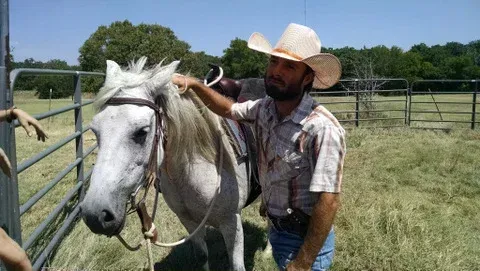 Cowboy in a hat leading a white horse in a grassy field on a sunny day