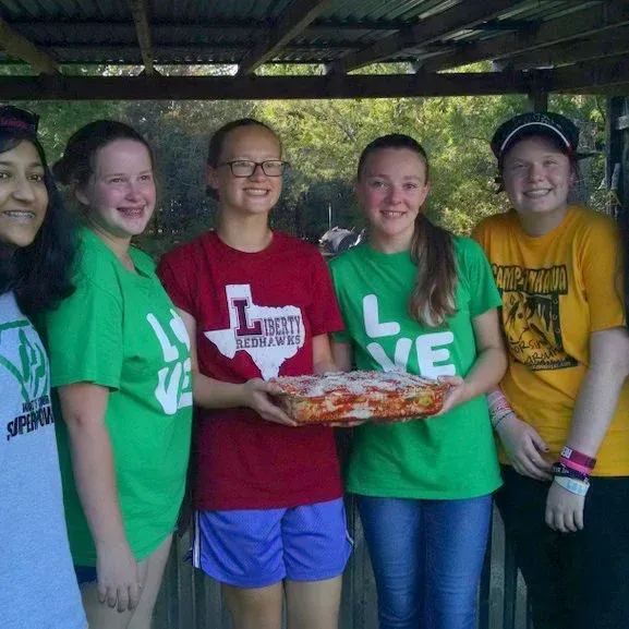 Five people in colorful shirts stand under a shelter, smiling and holding a tray of food.