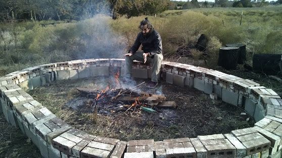Person tending a small fire in a circular stone fire pit outdoors with smoke rising.