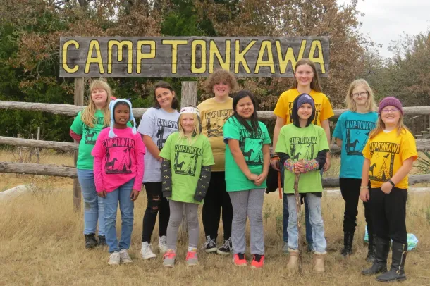 Group of campers posing in front of a Camp Tonkawa sign, wearing colorful shirts outdoors.
