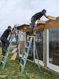 Two workers on ladders repairing a wooden roof frame outside a house