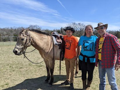 Three people standing beside a tan horse in a sunny pasture, all smiling at the camera.