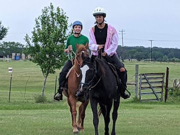 Two helmeted riders on horses in a grassy field near a gate and trees