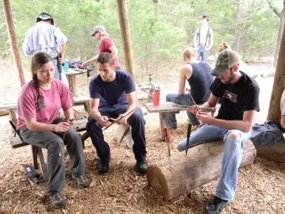 People sitting on logs outdoors, using phones and chatting in a wooded campsite.