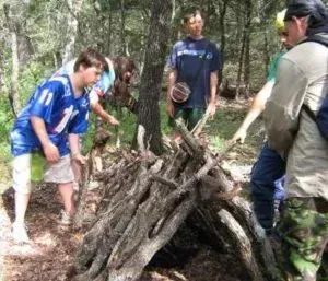 Three hikers in sportswear gather around a large pile of branches in a wooded area.