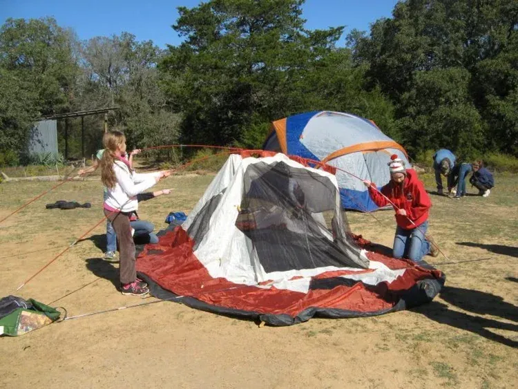 People setting up a red and gray camping tent in a sunny wooded area